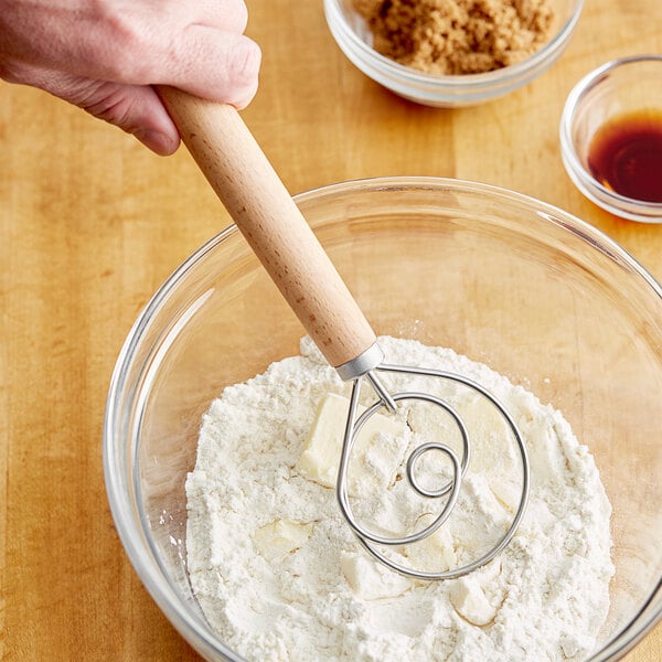 A hand mixing dough in a bowl with a Choice stainless steel Danish dough whisk with a wood handle.