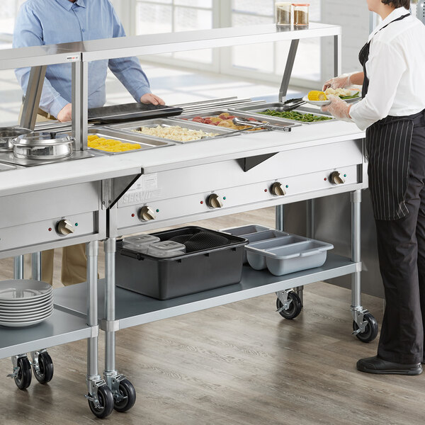 A man and woman using a ServIt electric steam table with sneeze guard in a kitchen.