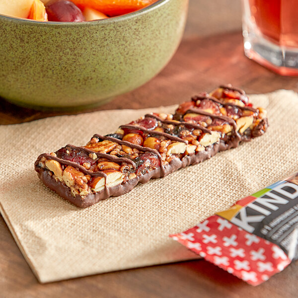 A KIND Dark Chocolate Cherry Cashew Bar with nuts and fruit on a napkin next to a bowl of fruit.