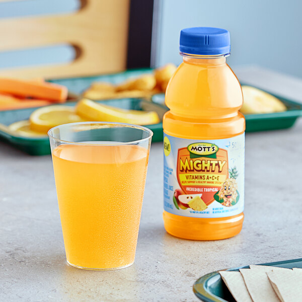 A bottle of Mott's Mighty Incredible Tropical Juice next to a glass of orange juice on a table.