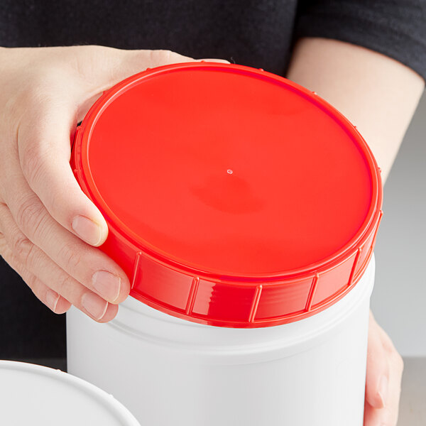 A hand holding a white plastic container with a red plastic lid.