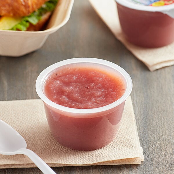 A plastic cup of Musselman's mixed berry applesauce on a table with a spoon and a sandwich.