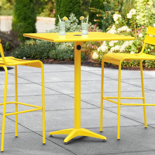 A Lancaster Table & Seating yellow powder-coated aluminum bar height table with two chairs on a patio.