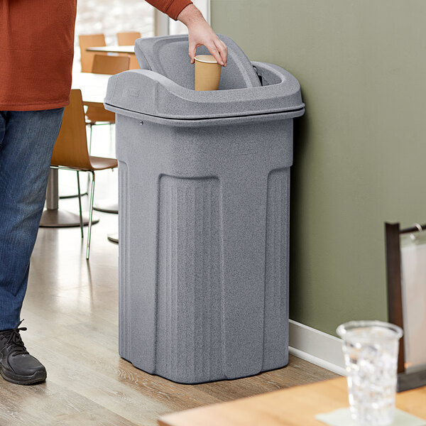 A man putting a paper cup in a Toter Slimline gray square trash can with a square lid.