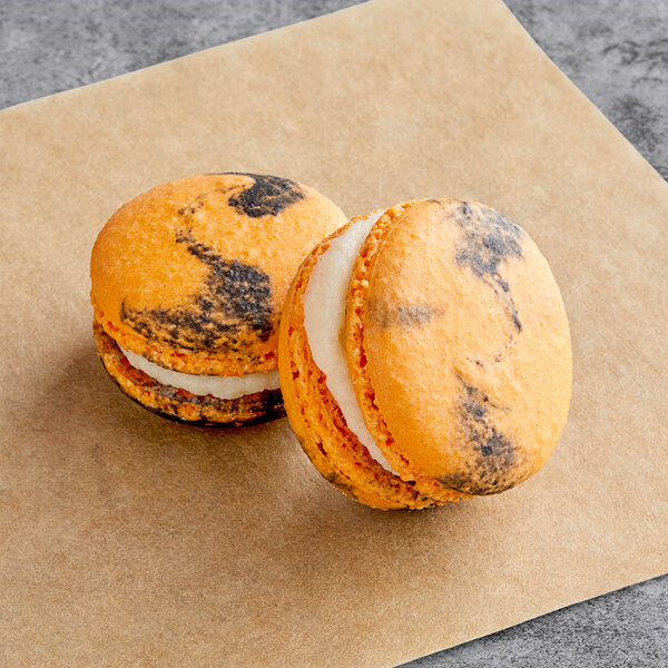 Two Macaron Centrale pumpkin cheesecake macarons with orange and black icing on a piece of paper.