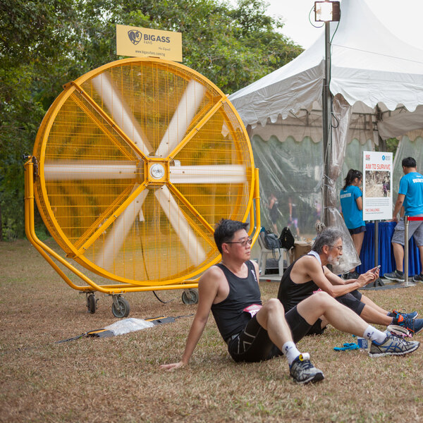 A large yellow Big Ass Fans AirGo portable floor fan in an outdoor catering setup with two men sitting on the ground.
