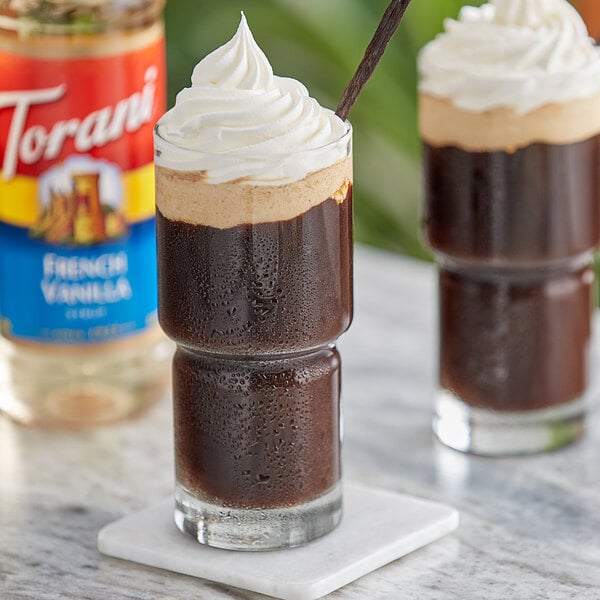 A glass of brown liquid with whipped cream on top next to a bottle of Torani French Vanilla Flavoring Syrup.