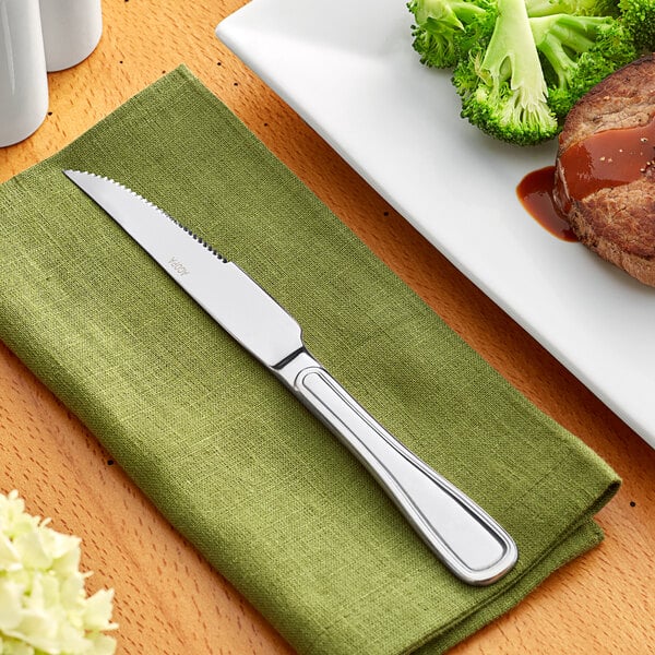 An Acopa Saxton stainless steel steak knife on a green napkin next to a plate of broccoli.