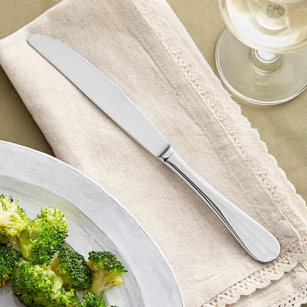 An Acopa stainless steel table knife on a white surface next to a plate of broccoli.