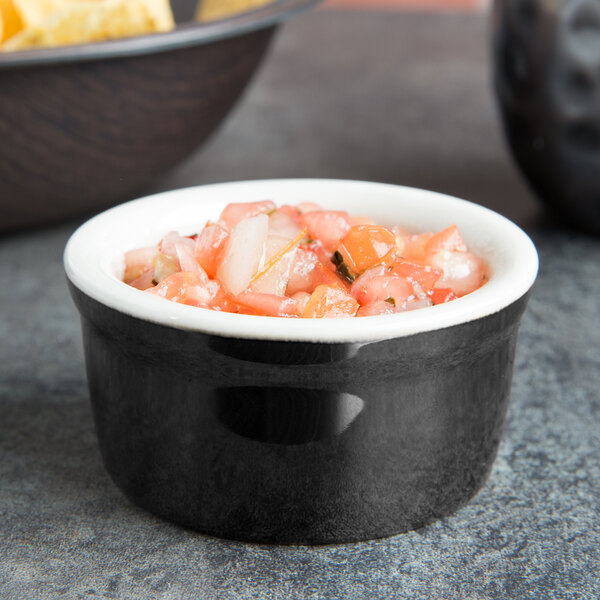 A black Tuxton ramekin filled with salsa next to a bowl of chips on a table.