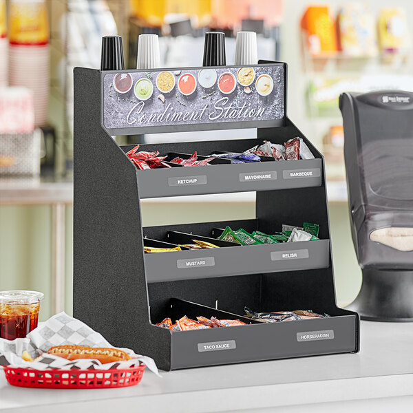 A black ServSense countertop condiment organizer with 12 sections and header decals on a counter.
