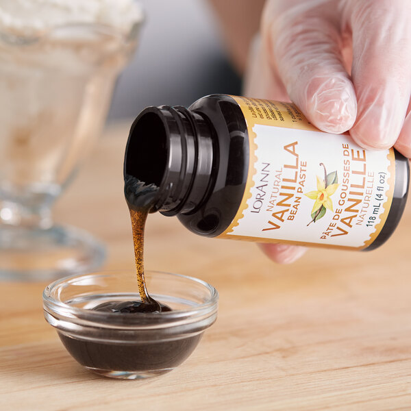 A gloved hand pours LorAnn Oils Natural Vanilla Bean Paste into a bowl of liquid.