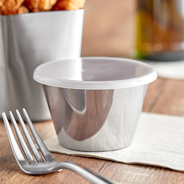 A close-up of a Tablecraft stainless steel sauce cup with a white lid on a table with a fork and a container of food.