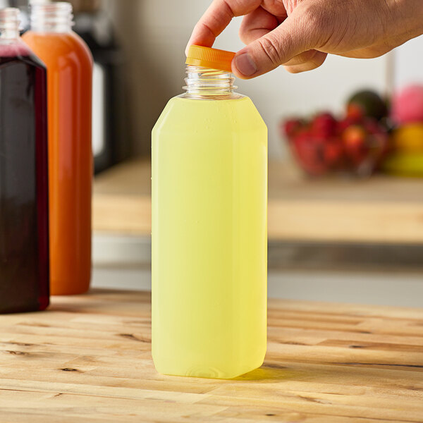 A hand holding a 32 oz. square PET clear juice bottle with yellow liquid and an orange lid.
