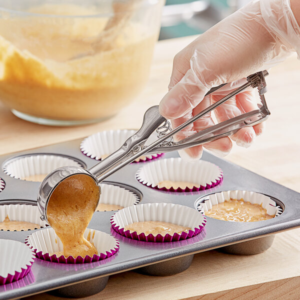 A person using a Choice stainless steel scoop to pour cupcake batter into a tin.