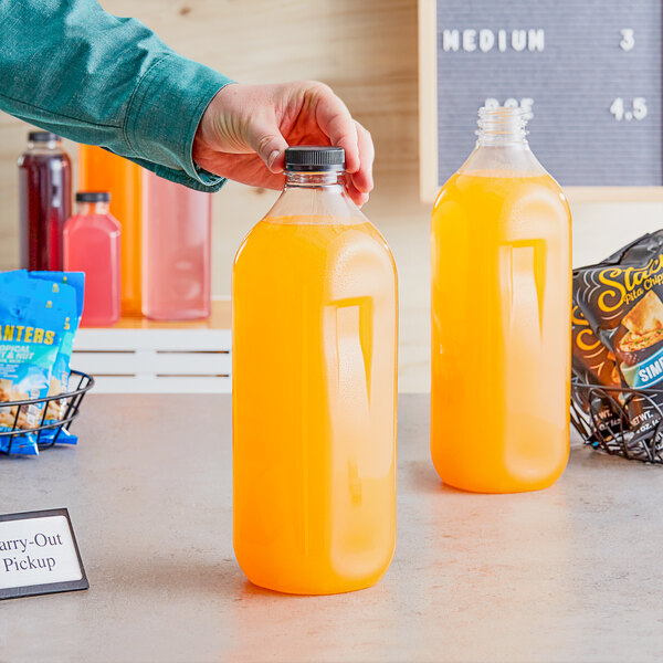 A hand holding a 59 oz. square clear bottle filled with orange liquid.