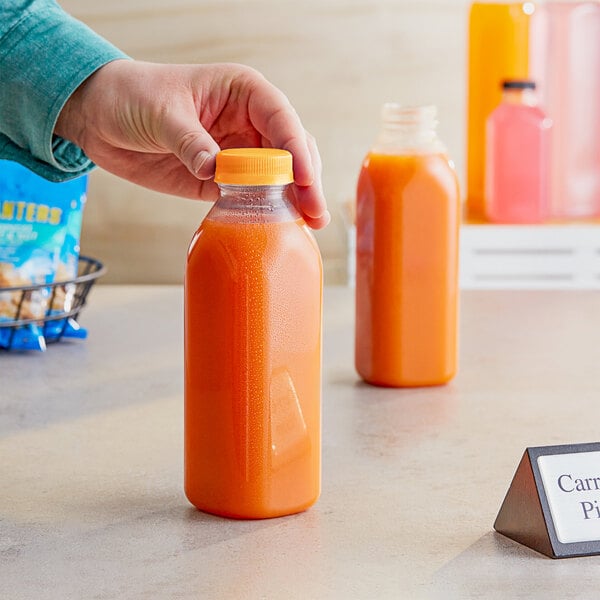 A hand holding a 16 oz. square PET clear juice bottle with orange liquid and a label on the counter.
