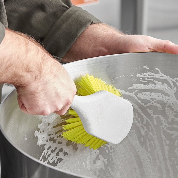A hand using a Lavex yellow pot scrub brush to clean a metal bowl.