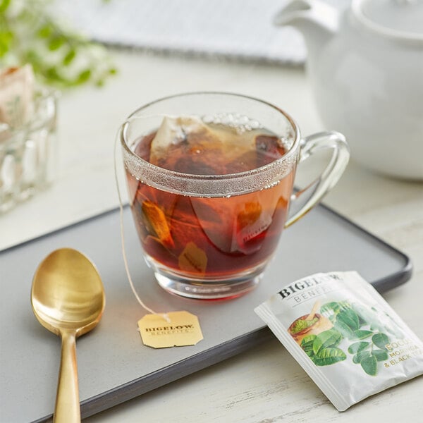 A glass cup of Bigelow Moringa and Black Tea with a tea bag and a teaspoon on a tray.