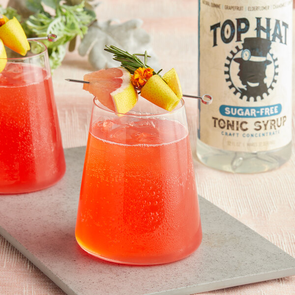 A bottle of Top Hat Provisions Sugar-Free Elderflower and Grapefruit Tonic syrup with glasses of red and orange drinks with fruit garnishes.