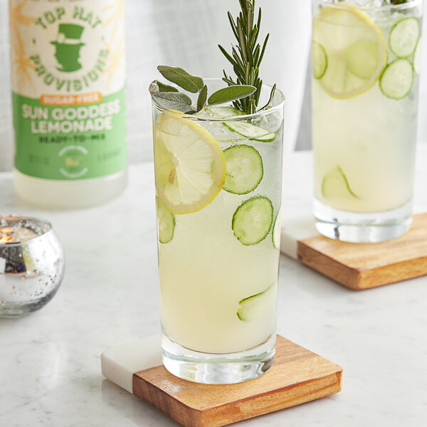 A glass of Top Hat Provisions Sun Goddess Cucumber Lemonade with lemons, cucumbers, and herbs.