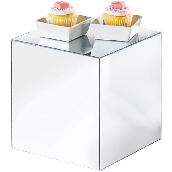 A white square container holding a cupcake with pink frosting on top placed on a mirror cube.