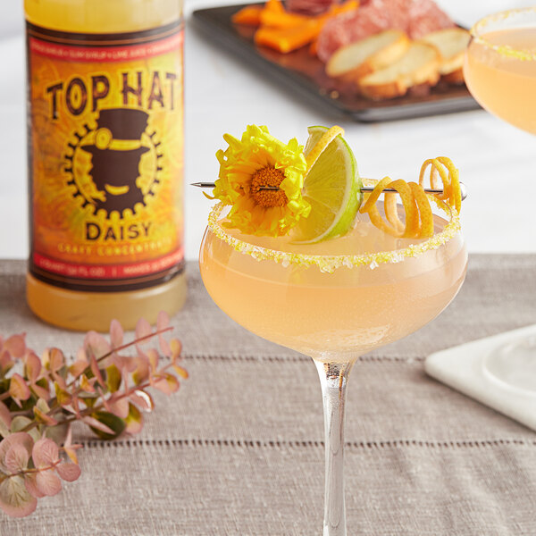 A glass of pink margarita with a yellow flower on top with a bottle of Top Hat Provisions Daisy and Margarita 5:1 Concentrate.