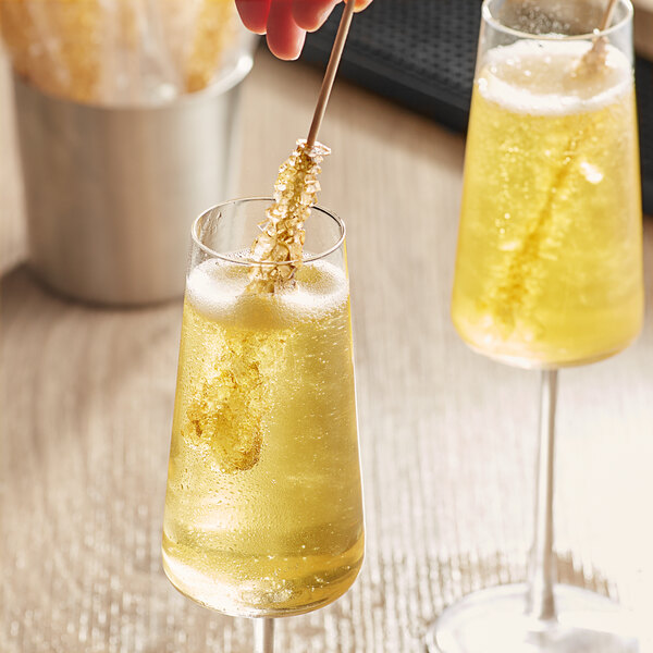 A hand holding a Roses Dryden and Palmer gold wrapped rock candy swizzle stick in a glass of yellow liquid.