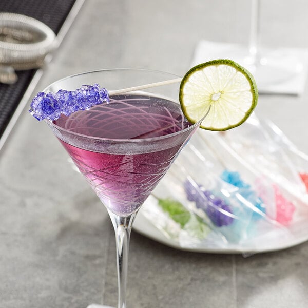 A lime slice on a Roses Dryden and Palmer purple rock candy swizzle stick in a glass of purple liquid.