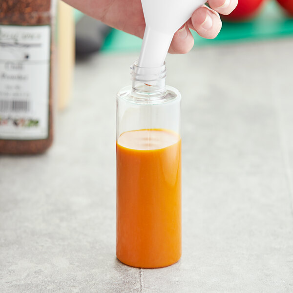 A hand pouring orange juice into a 4 oz. PET clear cylinder bottle using a white funnel.