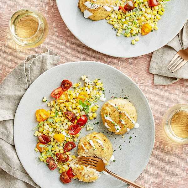 Two plates of Chesapeake Crab Connection blue crab cakes with corn and tomatoes on the table.