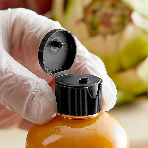 A hand in a glove holding a 24/410 black plastic flip top lid on a plastic bottle of yellow liquid.