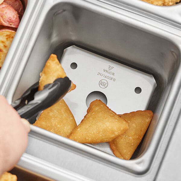 A hand using tongs to hold a stainless steel false bottom in a silver steam table pan.