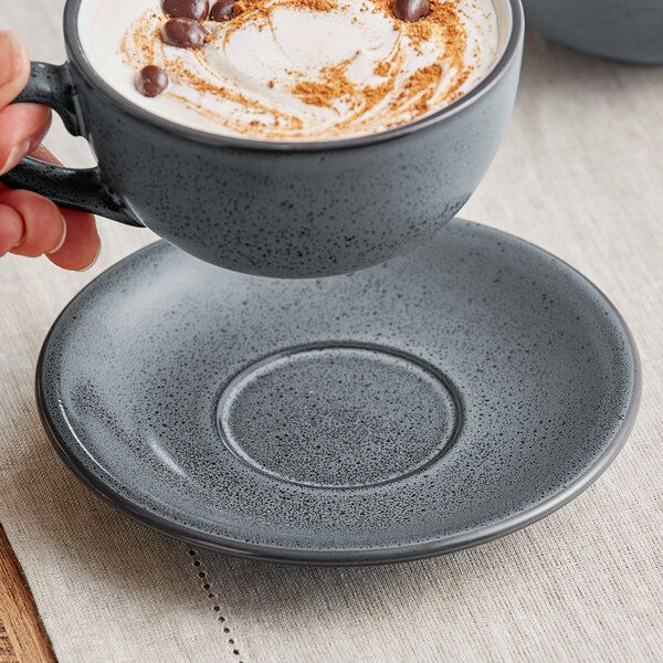 A hand holding a cup of coffee on a Midnight Blue Matte Acopa Embers stoneware saucer.
