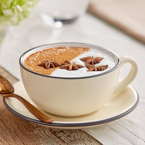 A cup of coffee with a white liquid and brown star anise in an Acopa Embers cream white stoneware cup on a saucer with a spoon.