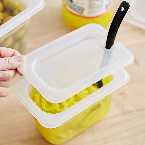 A hand using a black spoon to put olives in a translucent plastic container with a yellow lid.