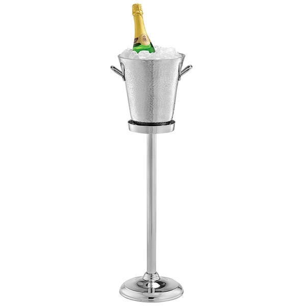 A Tablecraft stainless steel wine/champagne bucket stand with a champagne bottle in a bucket of ice on a table.