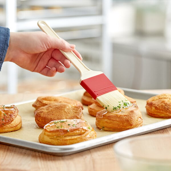 A hand using a Choice natural bristle pastry brush to spread pastry on a tray of pastries.