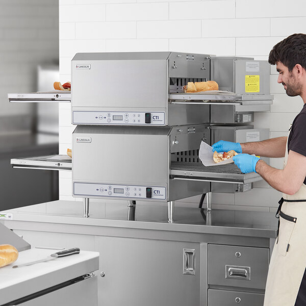 A man in a blue shirt and blue gloves using a Lincoln countertop conveyor oven to cook food in a professional kitchen.