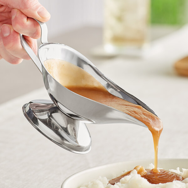 A person pouring gravy into a metal container using an American Metalcraft stainless steel gravy boat.