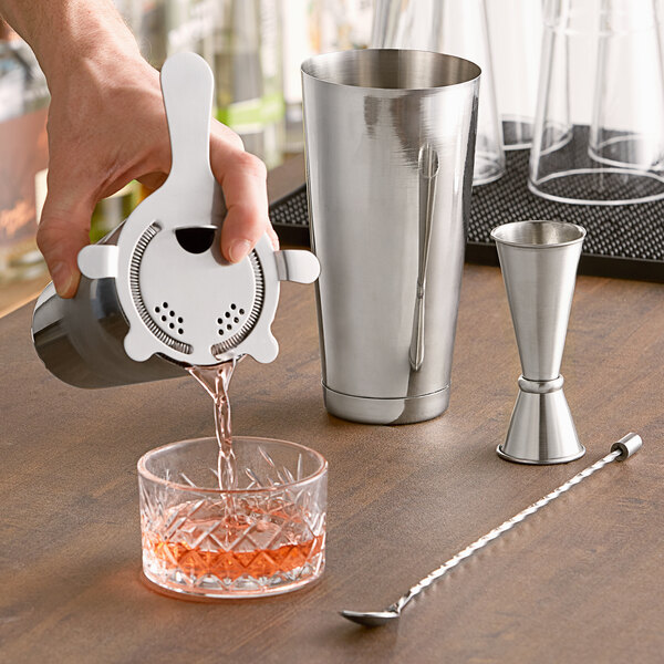 A person using an Acopa silver cocktail shaker to pour a drink into a glass.