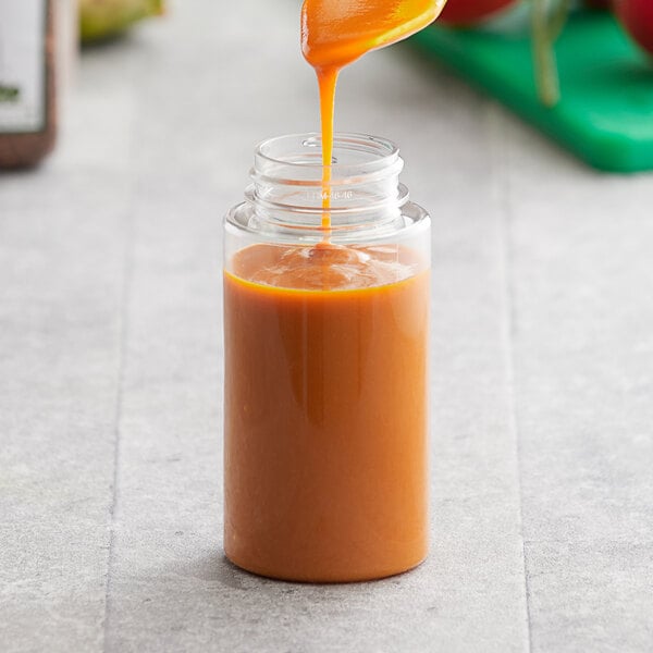 A spoon pouring caramel sauce into a clear plastic cylinder sauce bottle.