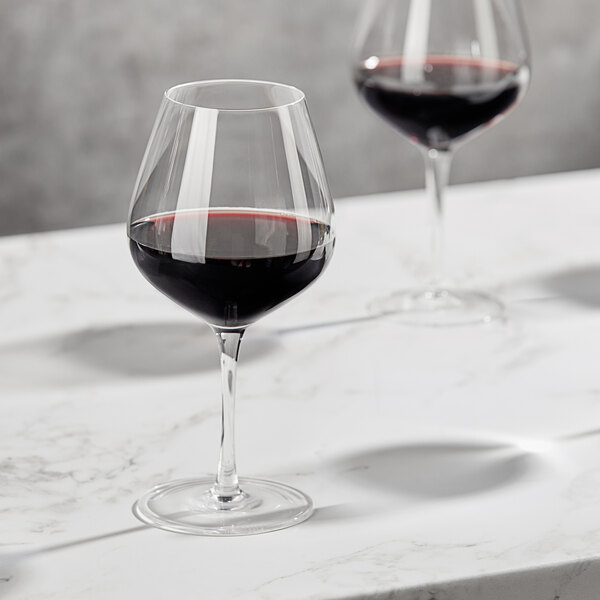 Two Della Luce Maia burgundy wine glasses on a marble table with red wine.