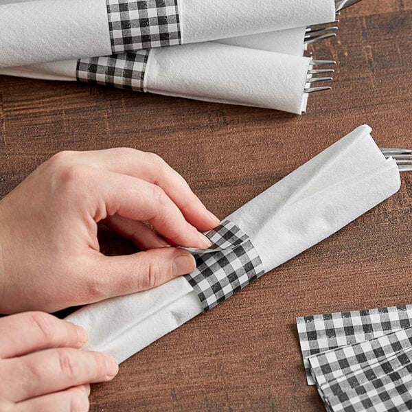 A person's hands folding a black and white checkered napkin with a Black Gingham Self-Adhering Paper Napkin Band.
