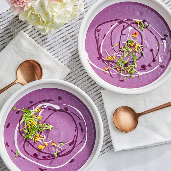 Two bowls of purple soup with white cream and green sprouts, spoons, and flowers.