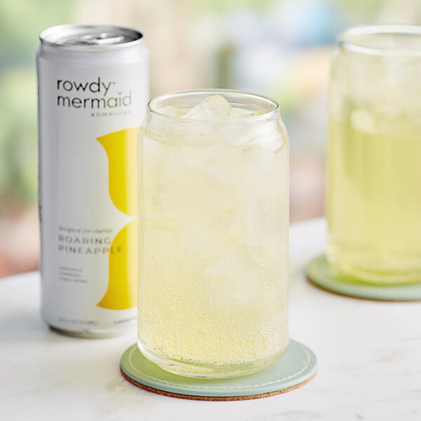 A close-up of a white and yellow Rowdy Mermaid Roaring Pineapple Kombucha can next to a glass of yellow liquid with ice.
