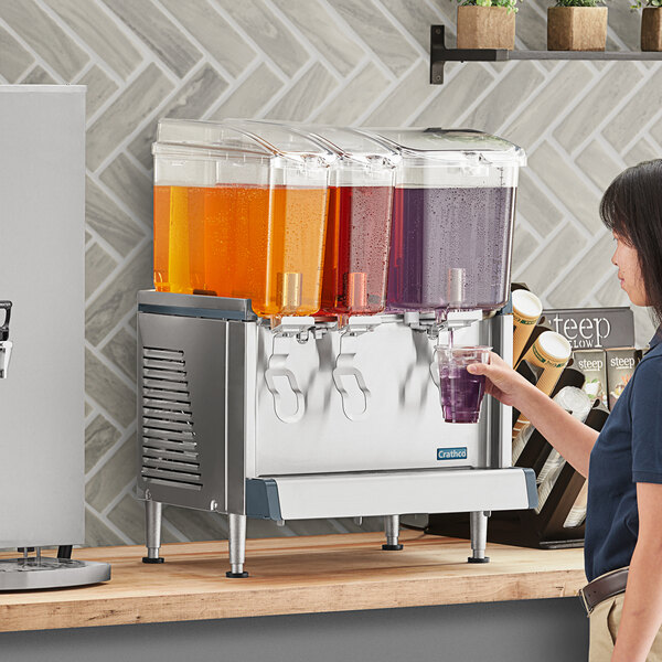 A woman using a Crathco triple bowl refrigerated beverage dispenser on a counter to pour a purple drink.