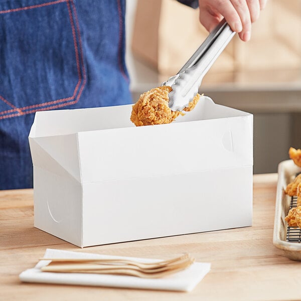 A person using tongs to put fried chicken in a white Choice lunch box.