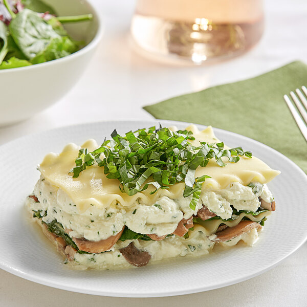 A white plate with a layer of Barilla Wavy Lasagna and a bowl of salad on a white surface.