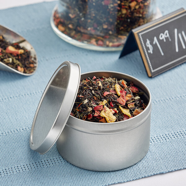 A silver deep tin with a lid full of tea leaves on a table with a spoon and a sign.